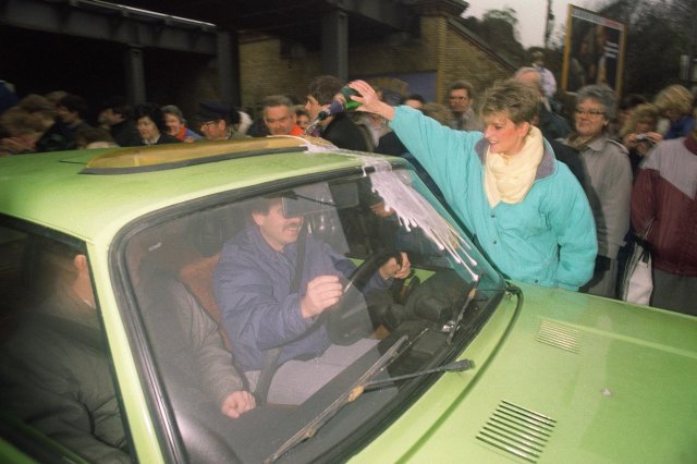 Mulher recebe moradores da Berlim Oriental enquanto  derrama Champagne em seu carro na Wollangstrasse, onde um novo ponto de passagem foi aberto em 13 de novembro de 1989