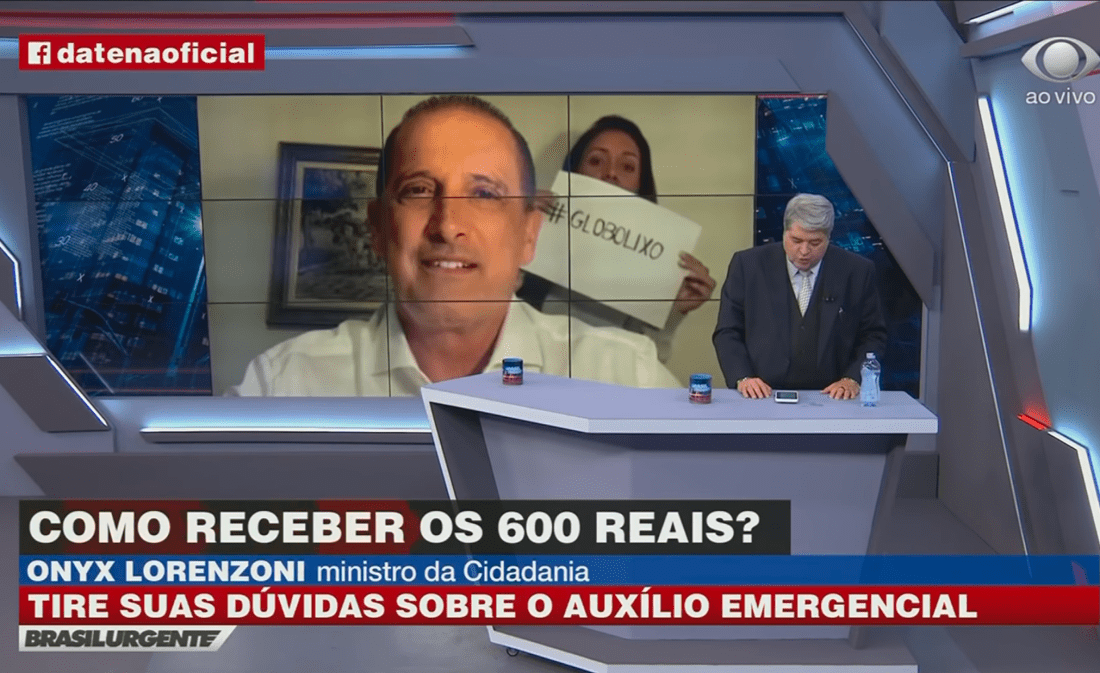 Mulher exibe cartaz “Globo Lixo” durante entrevista de Onyx Lorenzoni a Datena
