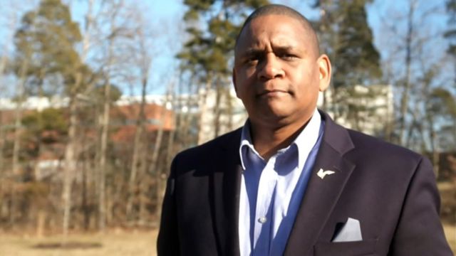 John Hollis, um homem negro de terno e com o cabelo curto, em uma paisagem ao ar livre